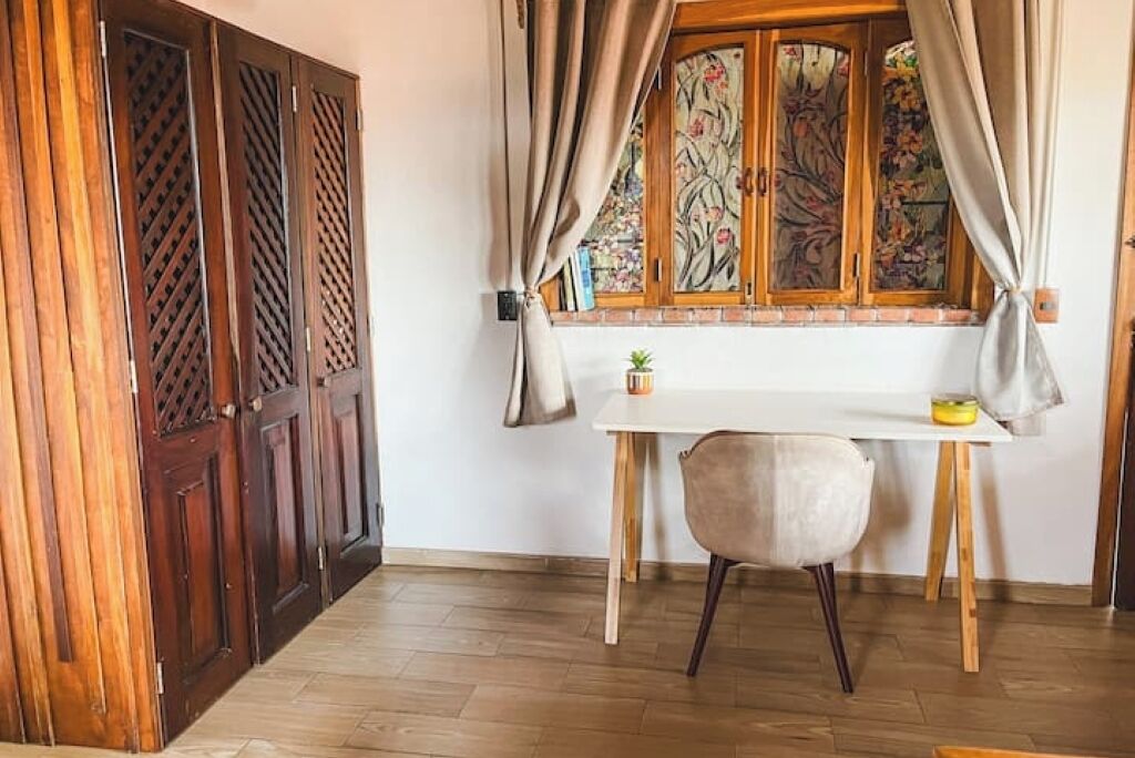Een kamer met een houten deur, een raam met bloemen gordijnen, een tafeltje en een stoel.