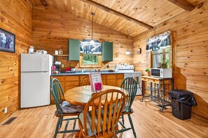 A wooden cabin interior with a dining table, chairs, a refrigerator, a microwave, and a window with a view of trees. - Pine View Cabin at Hocking Hills (South Bloomingville)