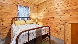 A wooden-paneled room with a bed, a window with a view of trees, and a wall-mounted lamp. - Pine View Cabin at Hocking Hills (South Bloomingville)