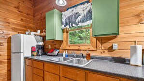 A kitchen with wooden cabinets, a refrigerator, a sink, and a window with a bear illustration. - Pine View Cabin at Hocking Hills (South Bloomingville)