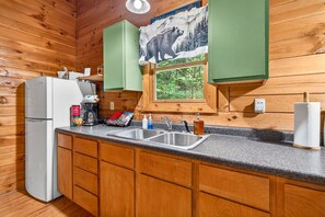 A kitchen with wooden cabinets, a refrigerator, a sink, and a window with a bear illustration. - Pine View Cabin at Hocking Hills (South Bloomingville)