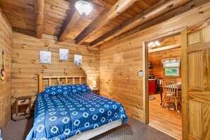 Ein Zimmer einer Holzhütte mit einem Bett, Holzwänden, einer Deckenleuchte und einer Tür zu einer Küchenzeile.