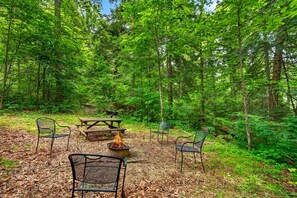 A forested campsite with picnic tables, metal chairs, and a campfire. - Wild Turkey Cabin at Hocking Hills (South Bloomingville)