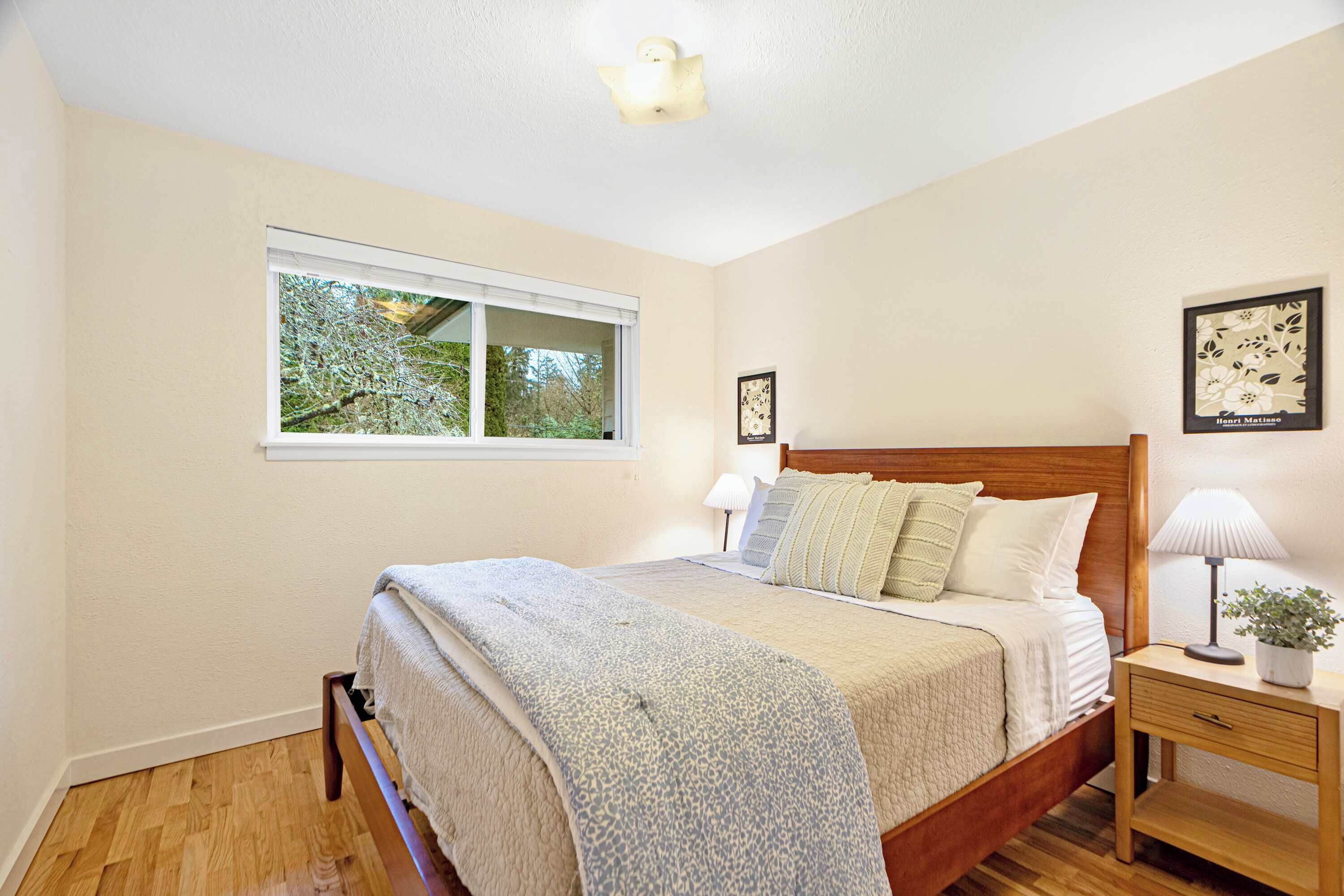 A bedroom with a wooden bed, white walls, a window, and a nightstand with a lamp.