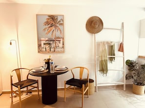 A dining area with a round table, two chairs, a framed picture of a palm tree, a straw hat on a ladder, and a potted plant. - Sea view 1-bedroom LUXURY and cosy apartment in beautiful Orient Bay with AC (Saint Martin)