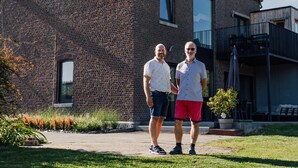 Two men standing in front of a brick building with large windows and a balcony. - Le Surdan (Bassenge)