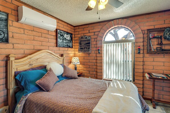 Una camera da letto con parete in mattoni, una testiera in legno, un letto con biancheria a motivi, una finestra con persiane e uno scaffale con libri e un casco.