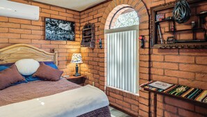 A room with a brick wall, a wooden bed with pillows, a nightstand with a lamp, a window with blinds, and a shelf with items. - Pool & Patio! 'Bike Haüs' By Catalina Mtn Trails (Tucson)