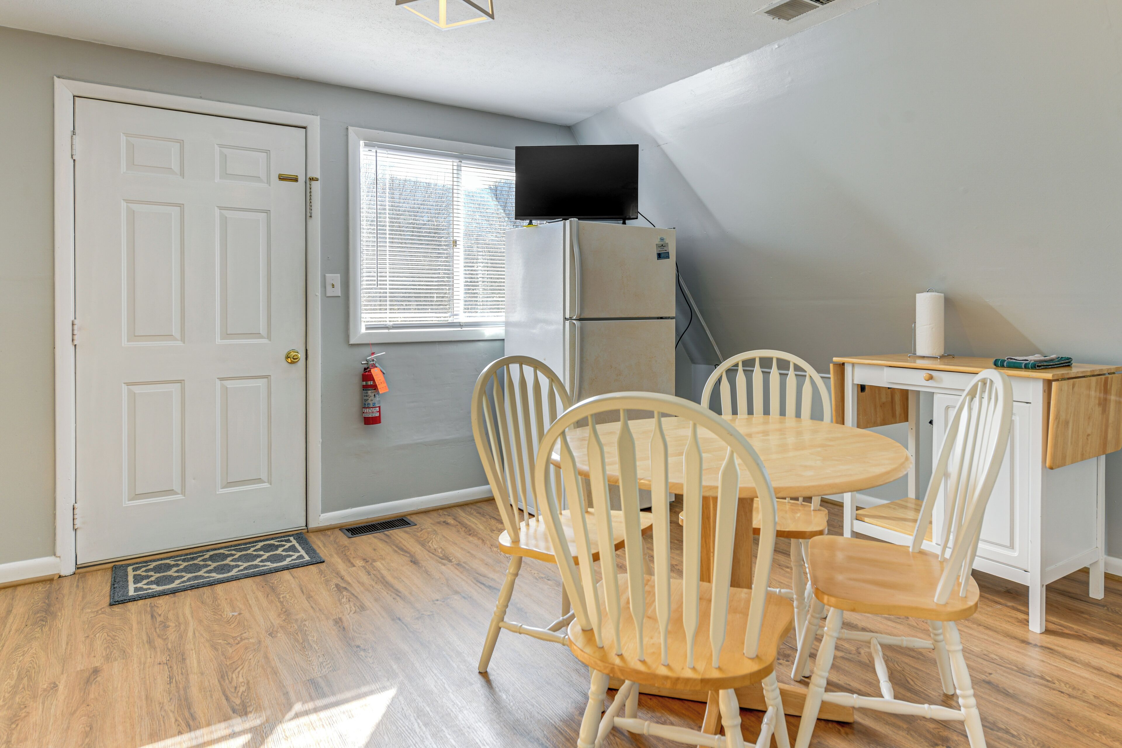 A compact kitchen with a dining table and chairs, a refrigerator, a flat-screen TV mounted on the wall, and a window with blinds.