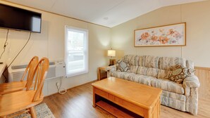 Ein Wohnzimmer mit einer Couch, einem Couchtisch, einem an der Wand montierten Fernseher, einem Fenster mit Jalousien und einem Gemälde an der Wand.