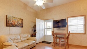 A living room with a beige leather sofa, a wooden dining table with two chairs, a flat-screen TV mounted on the wall, and a painting above the sofa. - Ski, Hike & Relax in the Smokies! Waynesville Apt (Waynesville)