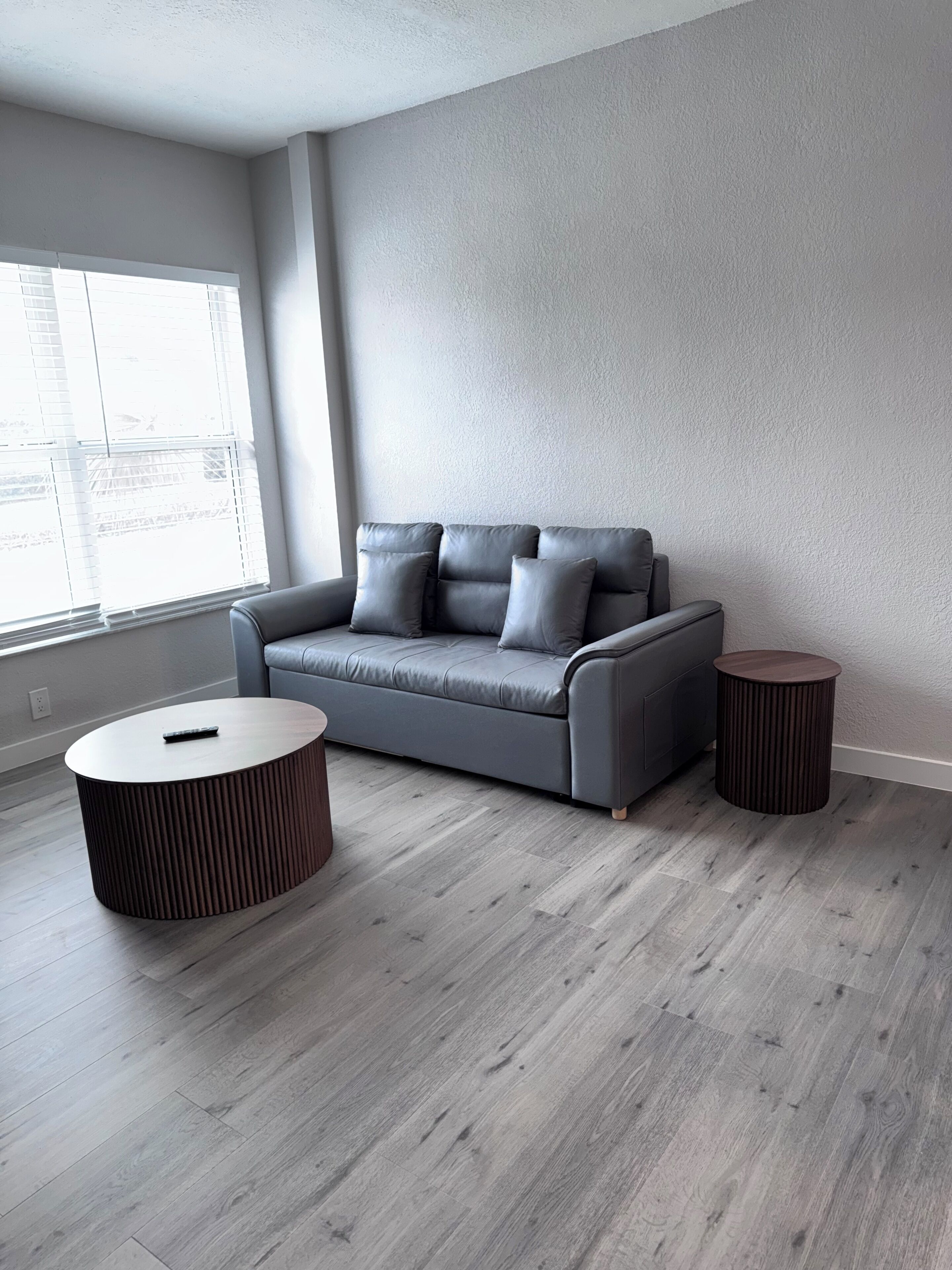 Un salon moderne avec un canapé gris, deux petites tables d’appoint rondes et une fenêtre avec des stores.