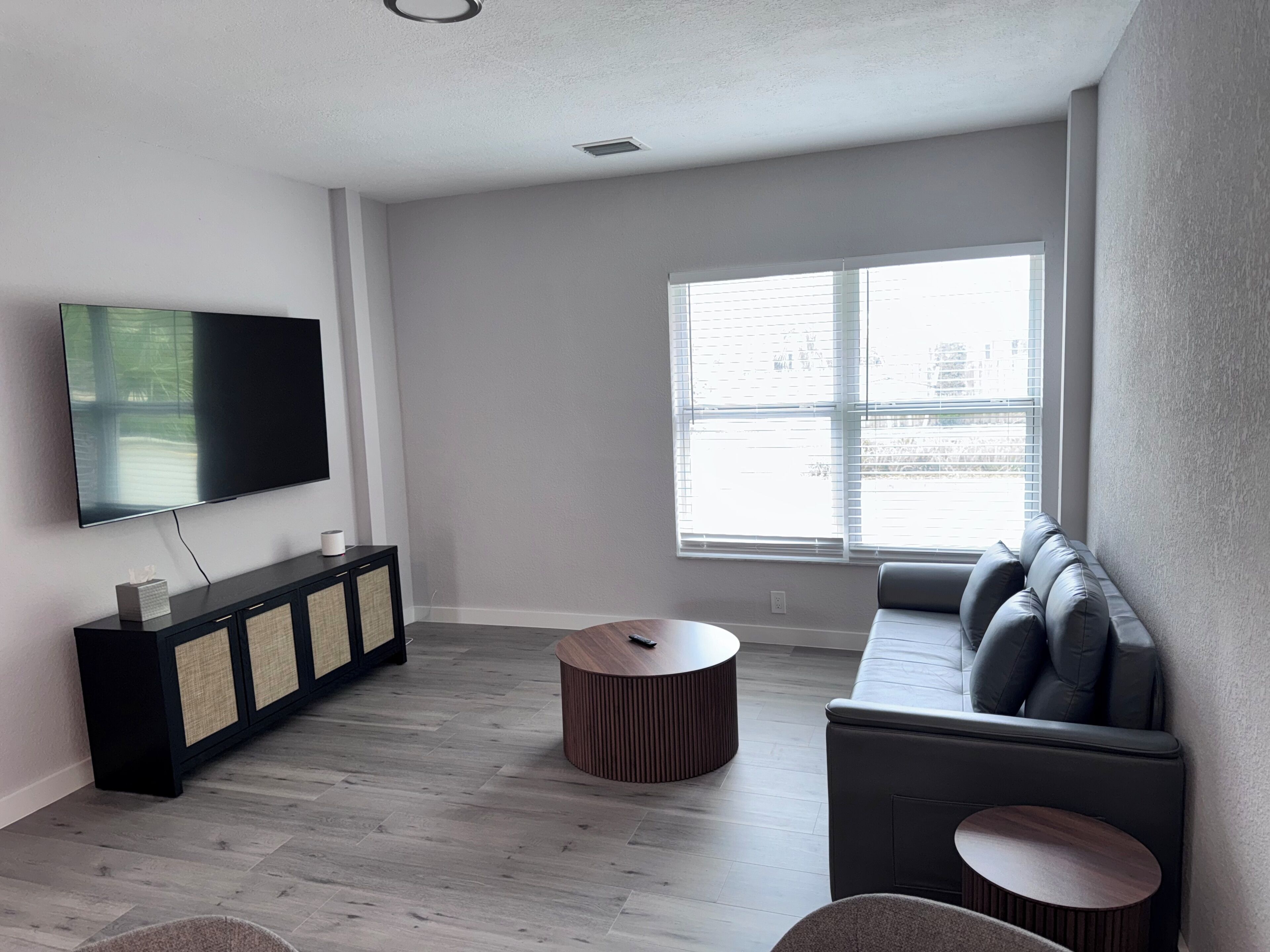 Ein modernes Wohnzimmer mit einem Flachbildfernseher, einer schwarzen Konsole, einem runden Couchtisch aus Holz, einem grauen Sofa mit Kissen und einem großen Fenster mit Jalousien.
