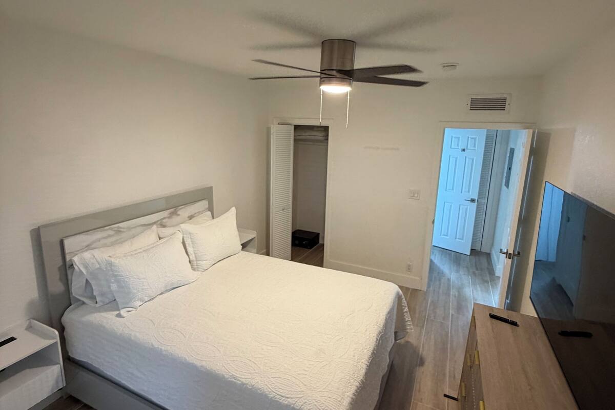 Une chambre à coucher comprenant un lit, un ventilateur de plafond, une table de chevet, une commode en bois et une porte donnant sur une autre pièce.