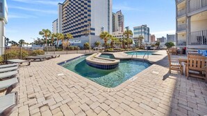 Ein Hotelpoolbereich mit einem Gebäude mit Schachmuster im Hintergrund, einem Pool mit kleinem Wasserfall und Liegestühlen rund um den Pool.