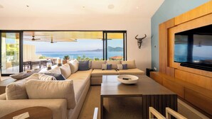 Un salon moderne avec un grand canapé, une table basse et un téléviseur fixé au mur. De grandes fenêtres offrent une vue sur la mer.