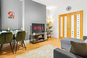 A modern living room with a flat-screen TV, a dining area with a table and chairs, and a large wooden door with glass panels. - Spacious 4 Bed Home | Free Parking & Fast WiFi (Denton)