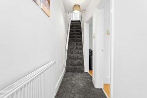 A narrow hallway with a carpeted staircase leading to a room with a white radiator and a light fixture above. - Spacious 4 Bed Home | Free Parking & Fast WiFi (Denton)