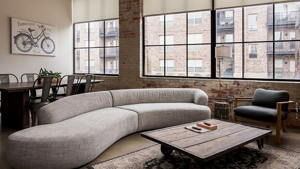 A modern living room with a large sectional sofa, a wooden coffee table, and a large window with a view of a brick building. - The SOHO Industrial Loft (Columbus)