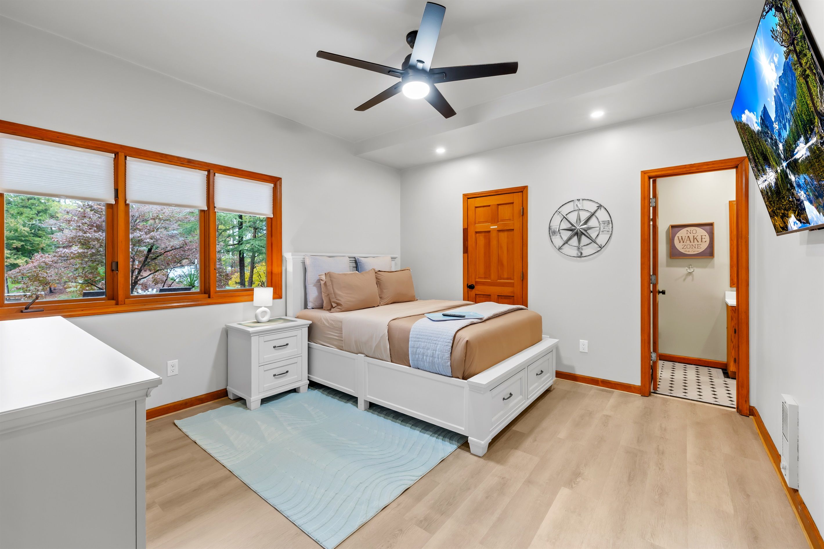 A bedroom with a bed, bedside tables, a ceiling fan, a window with blinds, a door, and a wall-mounted TV.
