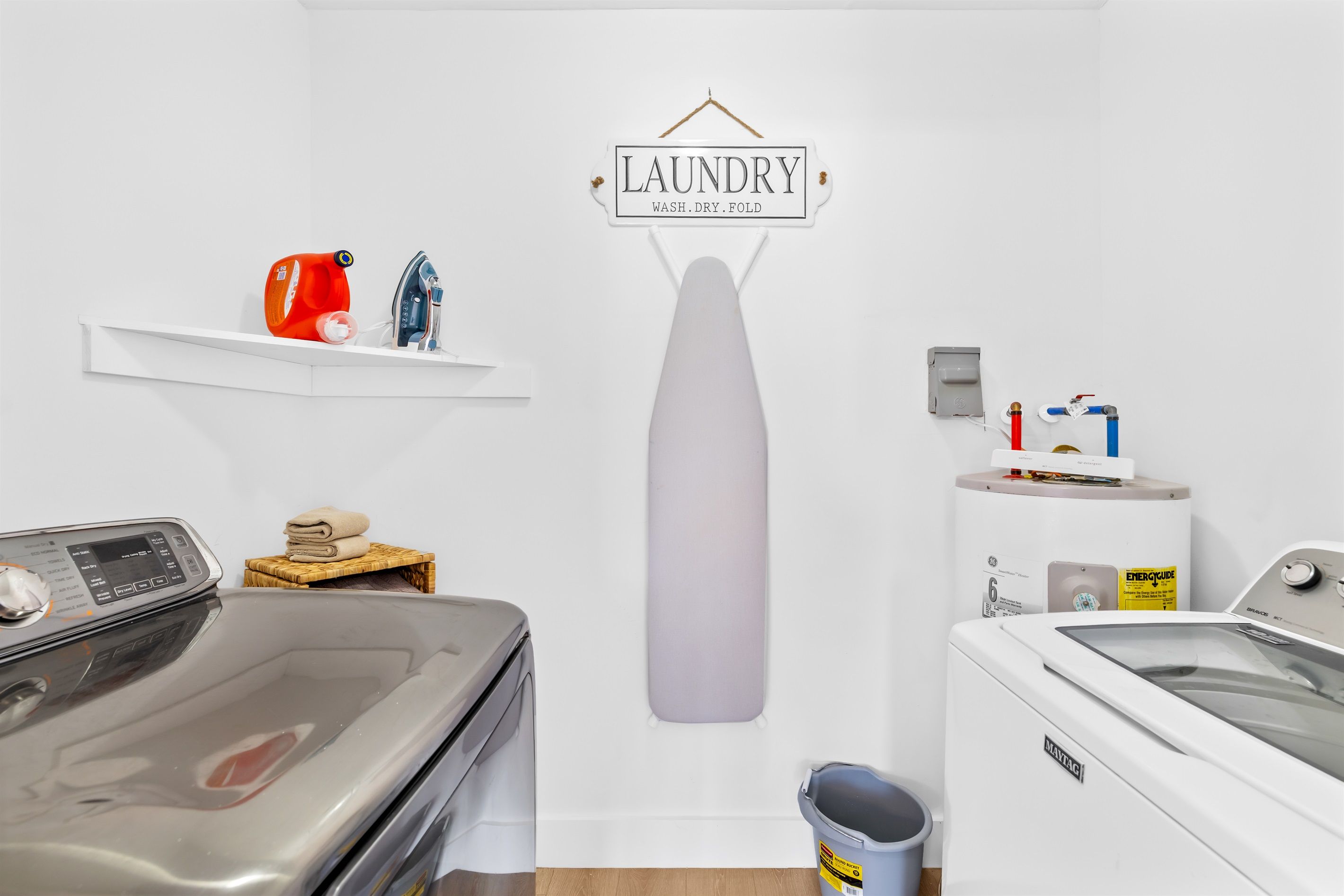 A laundry room with a washing machine, dryer, and ironing board. A sign above the ironing board reads "LAUNDRY WASH. DRY. FOLD."