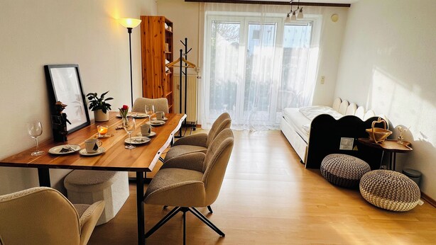 Un coin repas avec une table en bois dressée pour quatre personnes, un canapé avec des coussins, une petite table d’appoint et une fenêtre ornée de rideaux légers.
