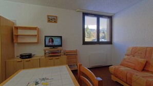 Ein Zimmer mit Fernseher auf einem Holztisch, einem Tisch mit Rautenmuster, einer Couch und einem Fenster mit Blick auf Bäume und Berge.