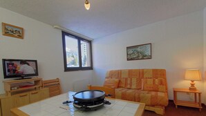 Ein kompakter Wohnraum mit Esstisch, Fernseher, Sofa und Fenster mit Blick auf umliegende Gebäude.