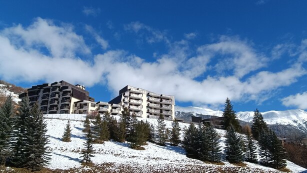 Ein mit Schnee bedeckter Berg mit einem modernen Gebäude, das mehrere Balkone und eine Skipiste im Vordergrund zeigt.
