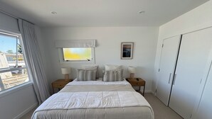 Une chambre avec un grand lit, deux tables de chevet avec des lampes, une fenêtre avec des rideaux, un tableau encadré au mur et une armoire.