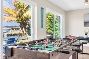 A foosball table in a room with large windows overlooking a body of water and palm trees. - BOAT LIFT! Waterfront Home w/ Hot Tub & Fire Pit! (Palm City)