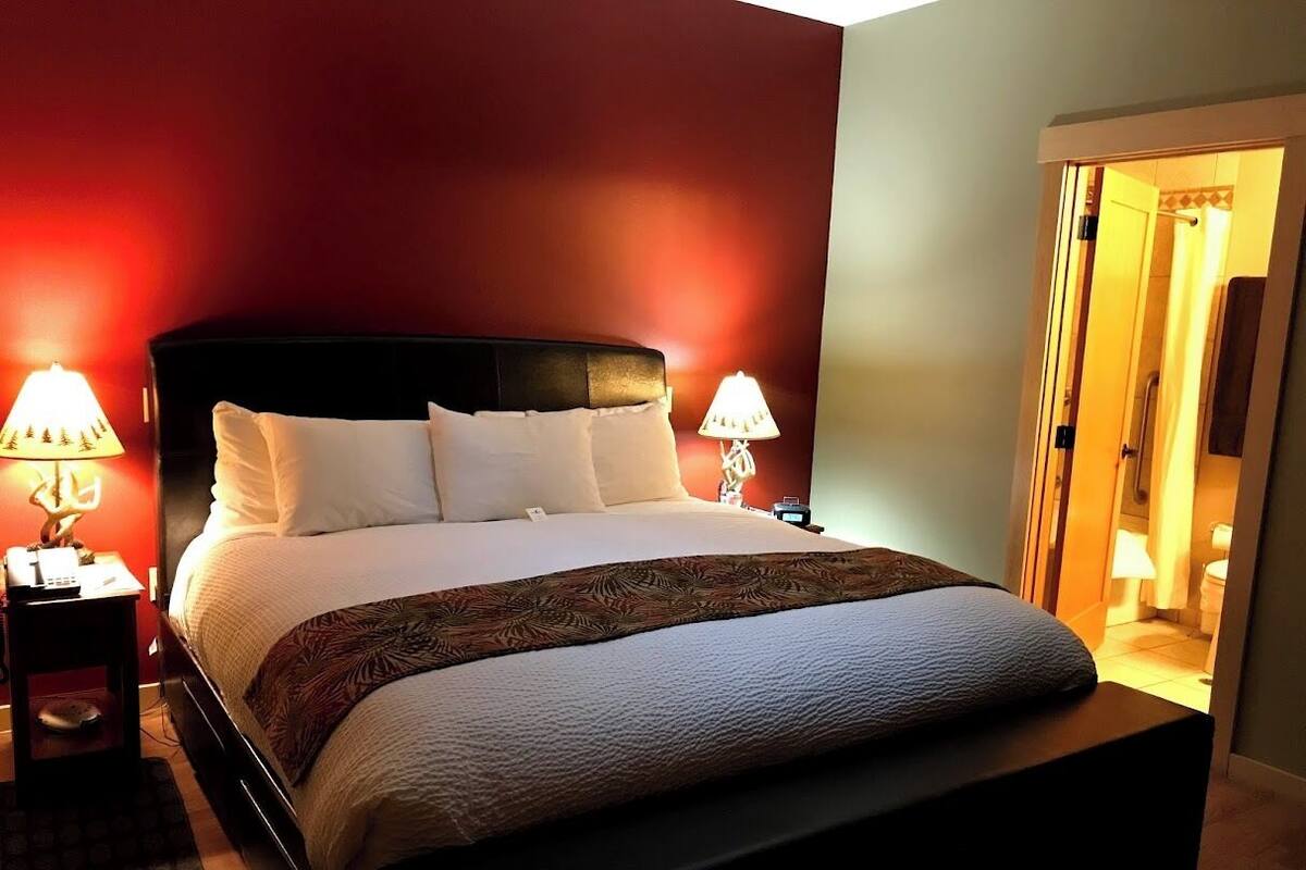A neatly made bed with a white comforter and a decorative throw blanket, flanked by two bedside tables with lamps, in a room with red walls and a visible bathroom door.