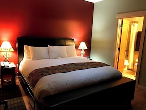 Una cama bien hecha con edredón blanco y manta decorativa, flanqueada por dos mesitas de noche con lámparas, en una habitación con paredes rojas y una puerta de baño visible.