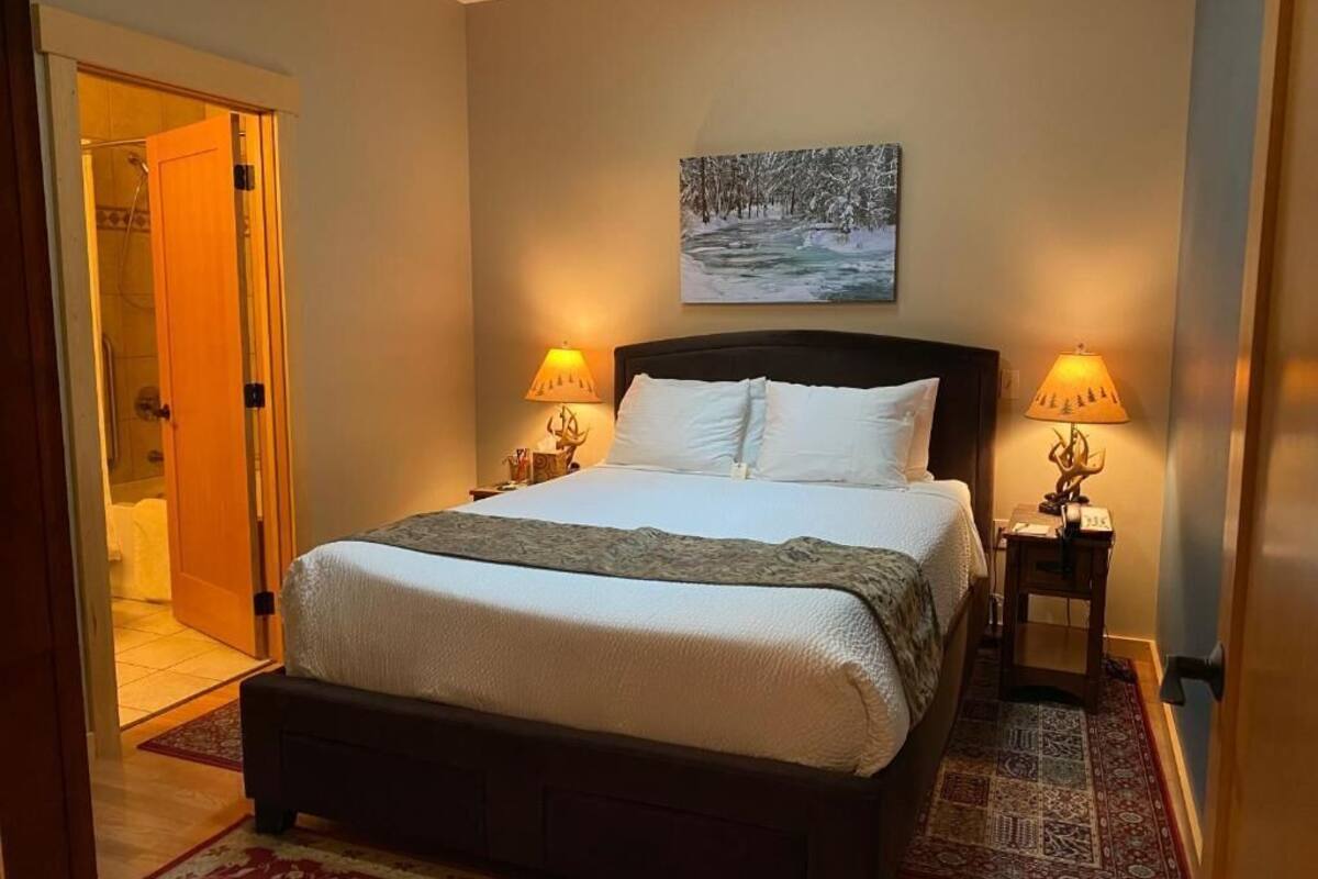 A neatly made bed with a white comforter and pillows, flanked by two bedside lamps. A painting of a snowy landscape hangs above the bed.
