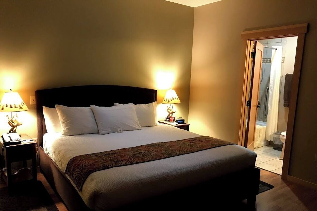 A well-lit hotel room with a large bed, two bedside lamps, a nightstand, and a view of the bathroom through an open door.