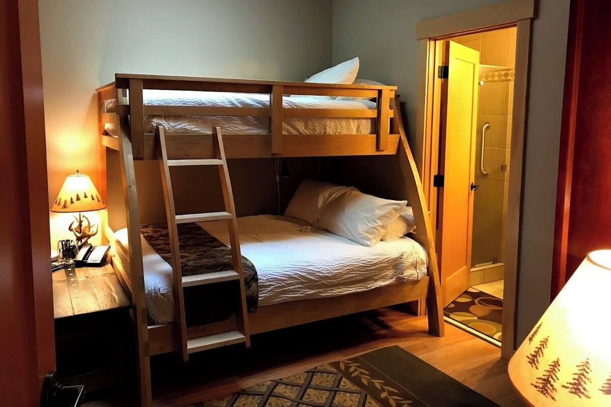 A bunk bed room with a ladder, a bedside table with a lamp, and a door leading to a bathroom.