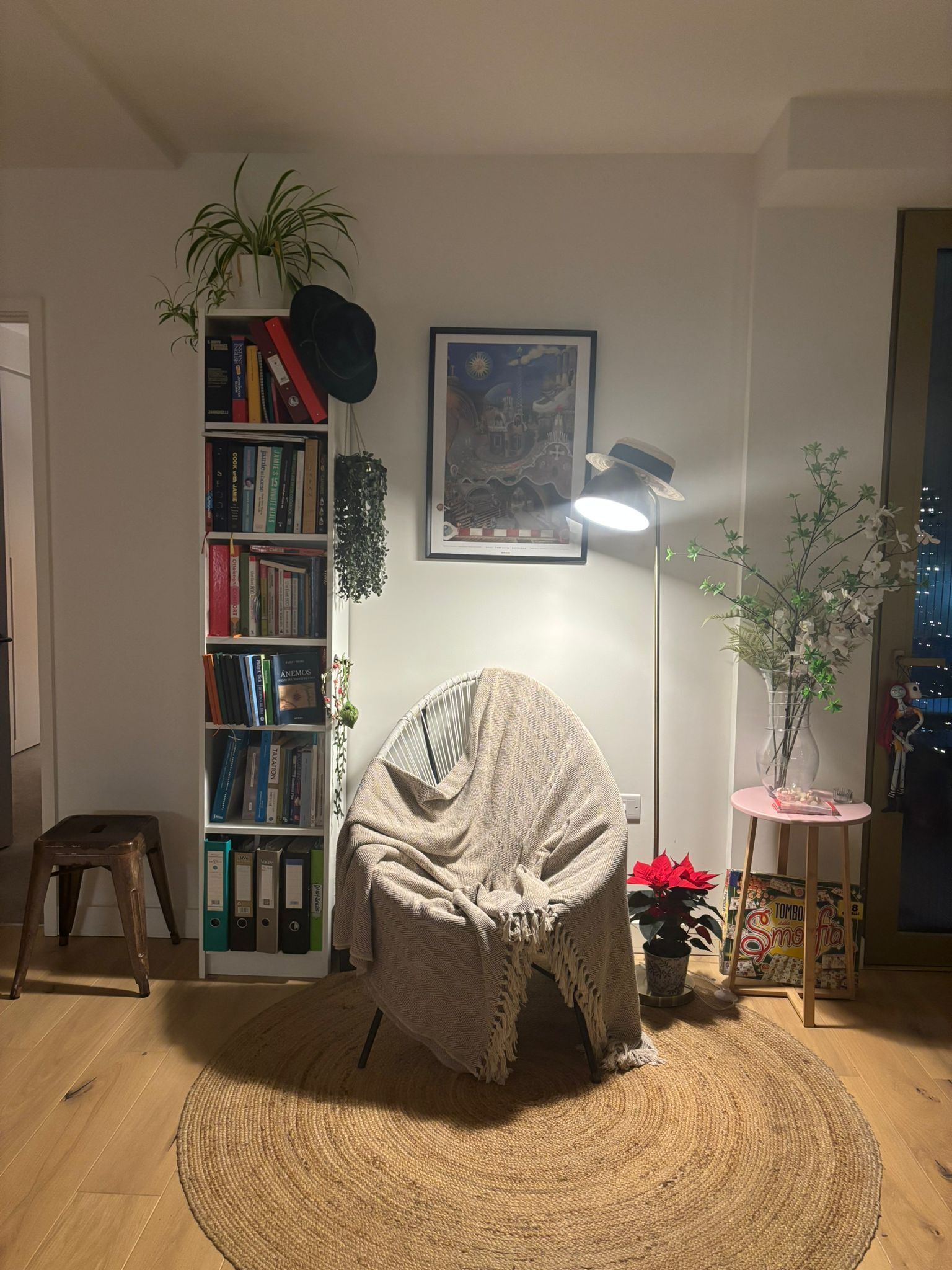 Un salon chaleureux avec un tapis rond en fibres naturelles, une bibliothèque, un tableau encadré, une lampe sur pied et une petite table sur laquelle est posé un vase de fleurs.