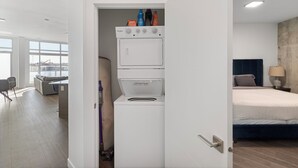 Une chambre d’hôtel compacte équipée d’un lave-linge, d’un lit, d’une table de chevet avec une lampe et offrant une vue sur l’extérieur grâce à de grandes fenêtres.