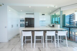 A modern kitchen with white cabinetry, a central island with bar stools, and a view of a cityscape through large windows. - 2 Bedroom (1 King & 1 Bunk Bed) Ocean View apartment (Main Beach)