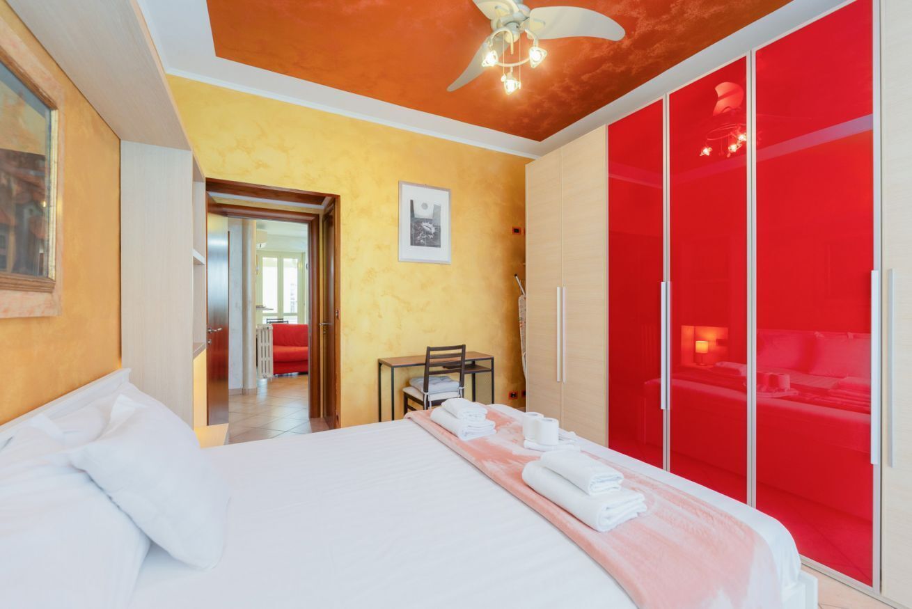 A hotel room with a bed, a red wardrobe, a ceiling fan, and a small table with a chair.