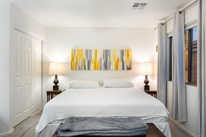 Ein Hotelzimmer mit einem Bett, zwei Nachttischlampen, einem großen abstrakten Gemälde und einem Fenster mit Vorhängen.