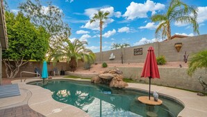 A swimming pool with a rock feature, surrounded by a patio area with a brick floor, palm trees, and a wall with decorative elements. - Desert Retreat w/ Pool, Trails & Mountain Views (Mesa)