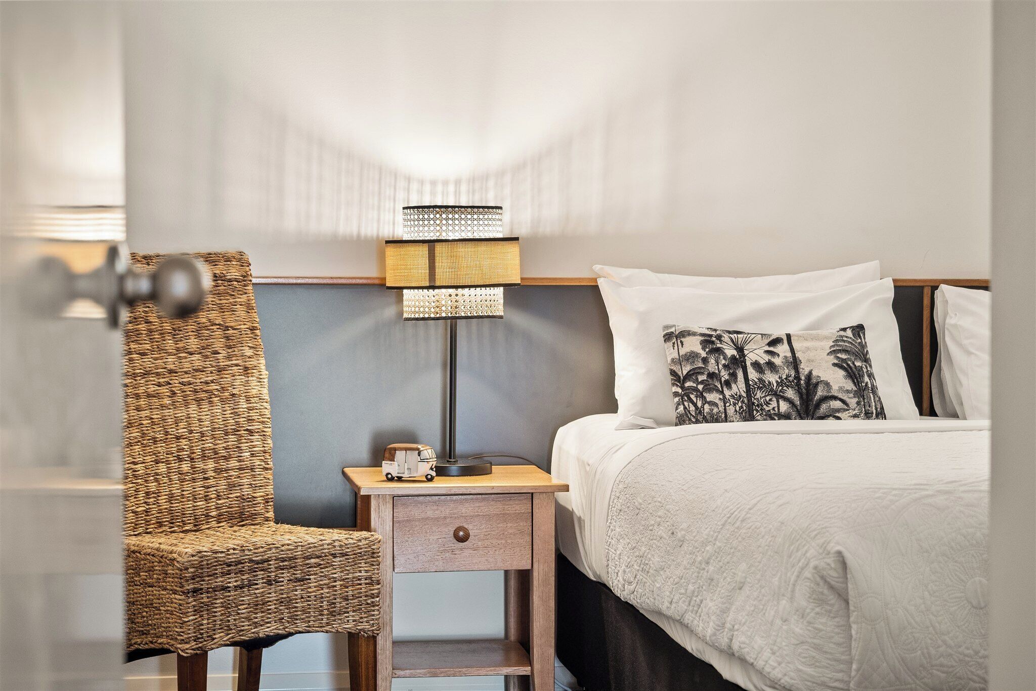 Una habitación de hotel con una silla de mimbre, una mesita de noche con lámpara, una cama con ropa blanca y una almohada decorativa.