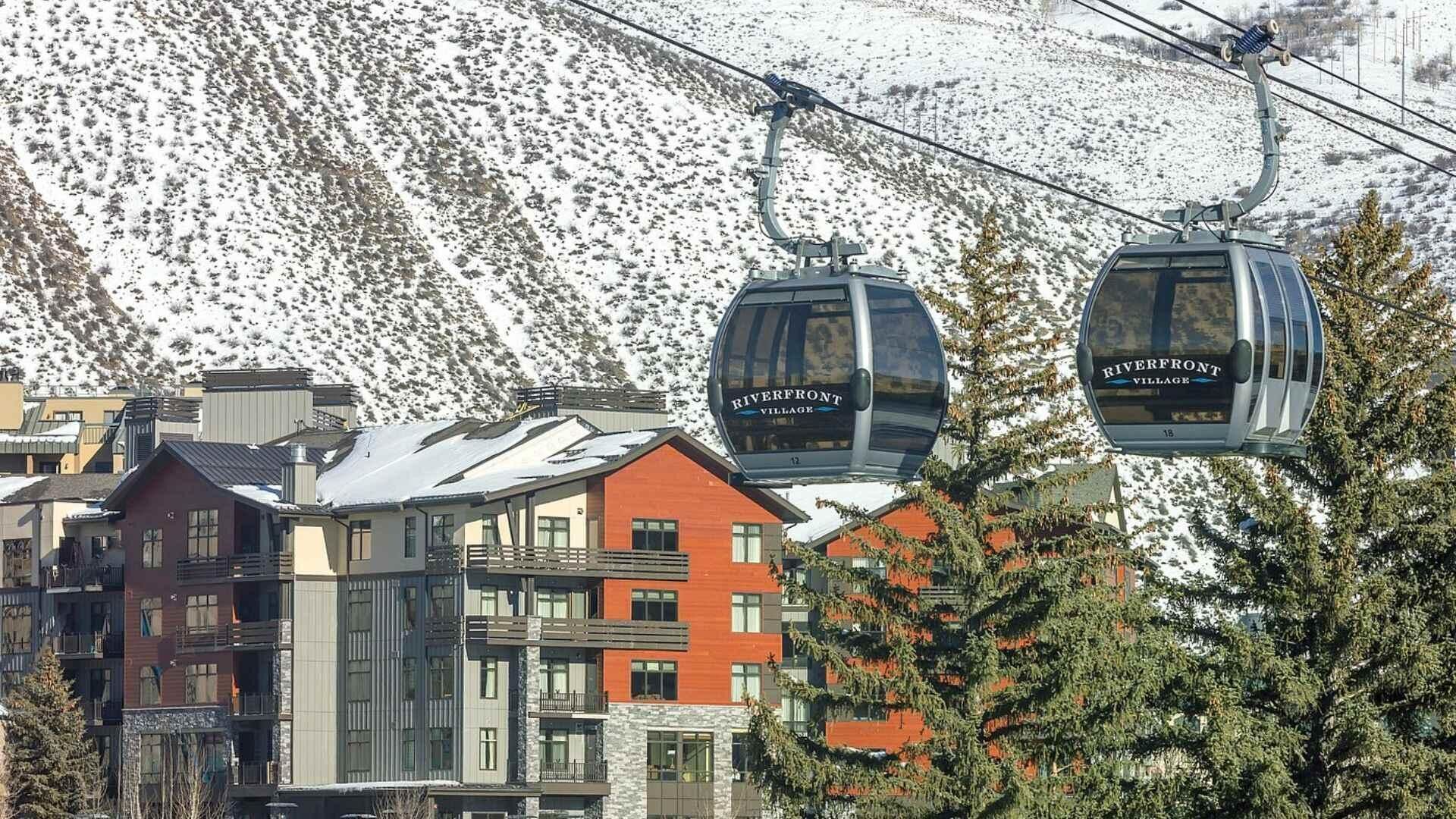 A ski lift with cabins labeled "Riverfront Village" against a backdrop of snow-covered mountains and buildings.