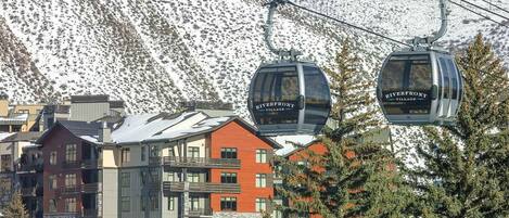 A ski lift with cabins labeled "Riverfront Village" against a backdrop of snow-covered mountains and buildings.