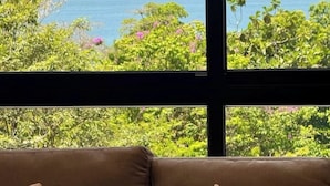 Ein Zimmer mit großem Fenster und Blick auf das Meer und üppige Vegetation.