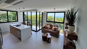 Eine moderne Küche mit Kochinsel, Essbereich inklusive Sofa und großen Fenstern mit Blick auf Grünflächen und das Meer.