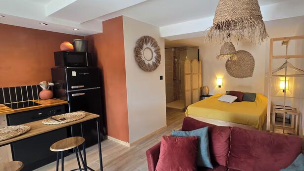 Habitación de hotel moderna con kitchenette, una cama con colcha amarilla, una maceta colgante y una silla de madera.