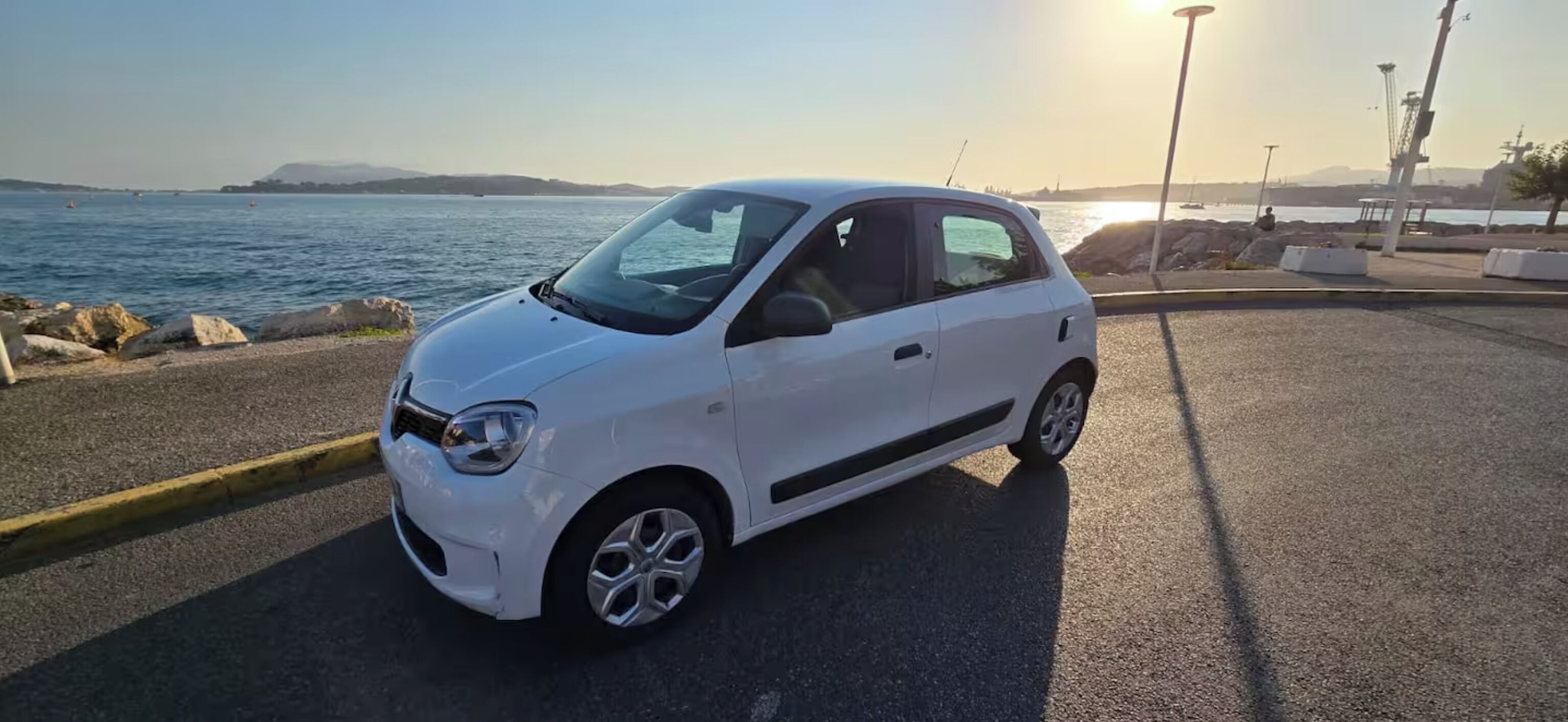 Une petite voiture blanche est garée sur une surface pavée, près d’une étendue d’eau, sous un ciel dégagé.