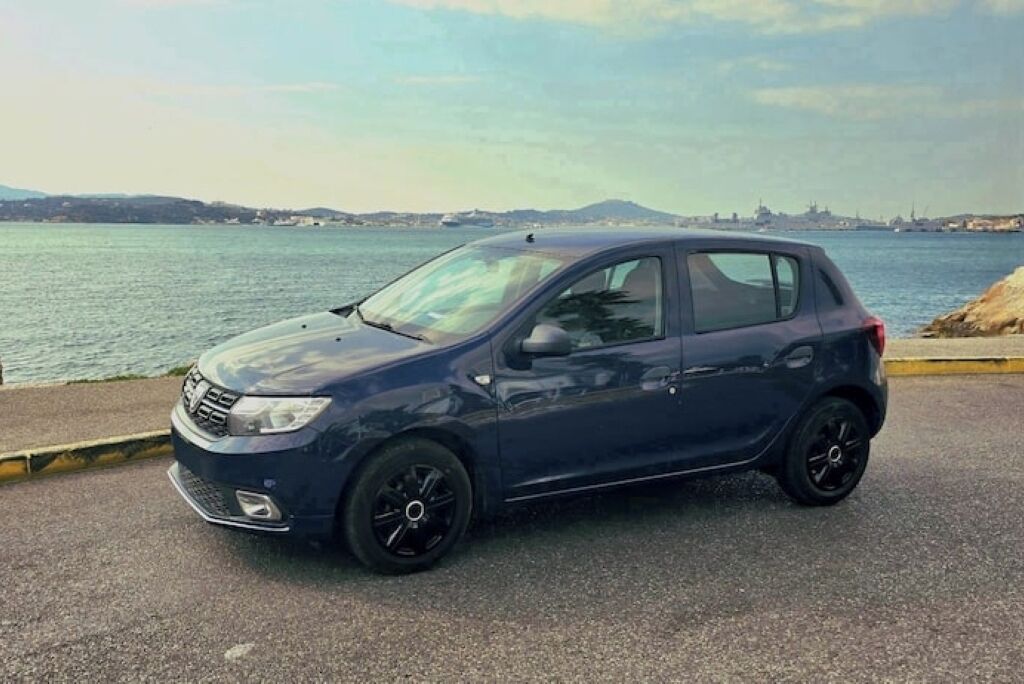 A dark blue car parked on a coastal road with a view of the sea and distant hills.